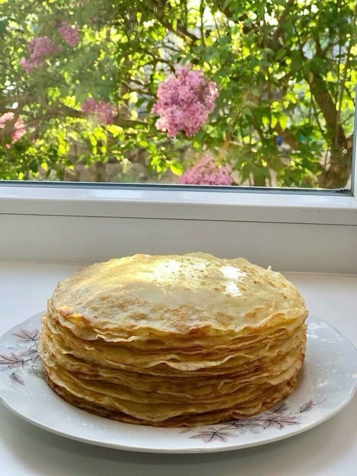 Stack of traditional Eastern European pancakes on a plate by a window with blooming lilac flowers outside.