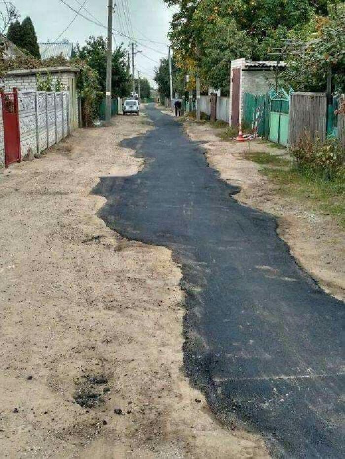 Uneven patch of asphalt covers a narrow dirt road in a quiet residential area, reflecting Eastern Europe road repair humor.