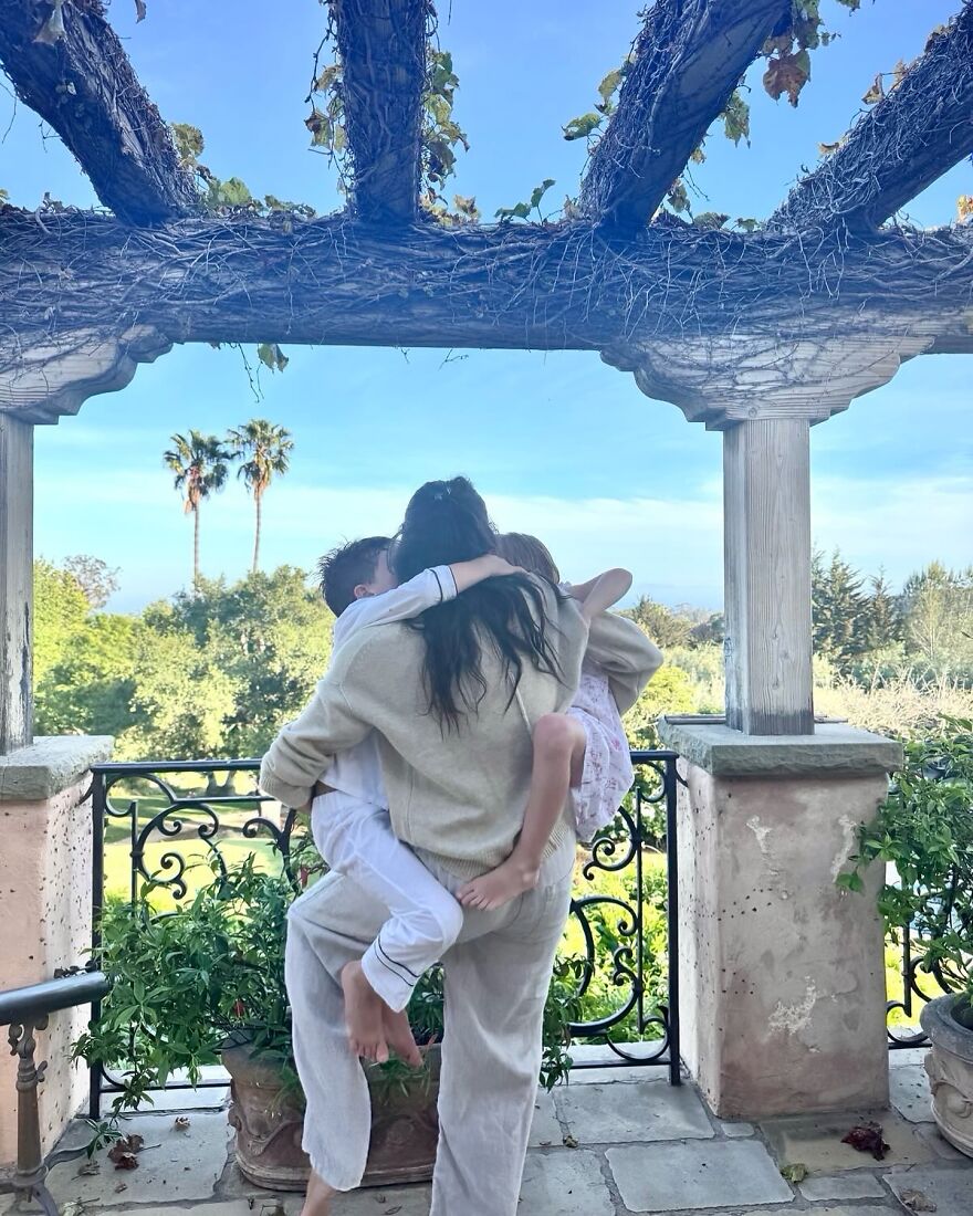 Meghan Markle carrying her kids outdoors under a pergola with greenery and palm trees in the background.