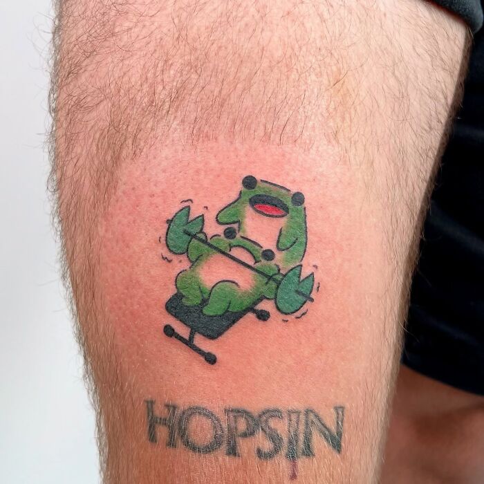 Tattoo of an adorable frog lifting weights on a person's thigh, showcasing animal tattoos by Korean artist Buoy.