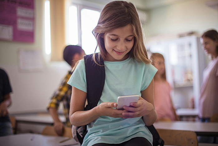 Young girl using smartphone in classroom while a sister accuses favoring nephew and niece in background discussion.