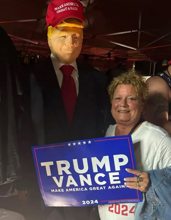 A woman posing next to a Donald Trump figure holding a Trump Vance 2024 campaign sign in a dimly lit setting.