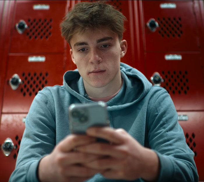 Teen boy looking at phone with red lockers behind, representing teen girl and boyfriend tormented by sick catfish online.