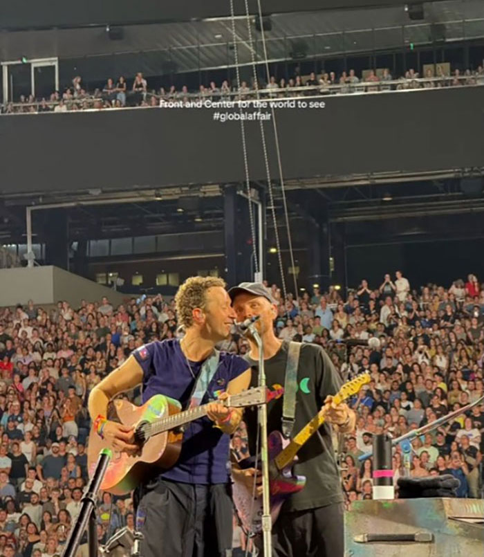 Coldplay band members performing on stage at a concert with a large crowd in the background. Coldplay band members performing on stage at a concert with a large crowd in the background.