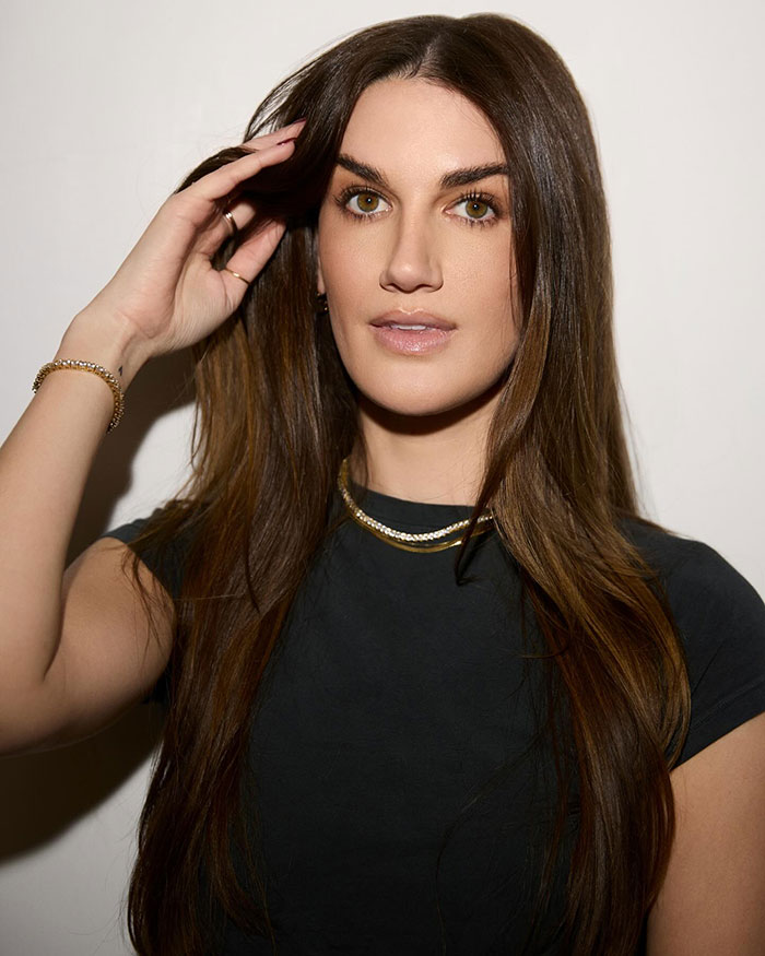 Woman with long brown hair touching her hair, wearing gold jewelry and a black shirt, related to celebrity facialist disfigurement claims.