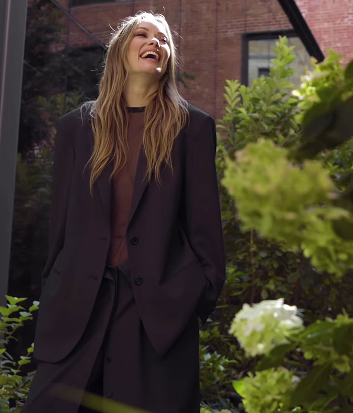 Olivia Wilde wearing a sheer outfit and flashing her toned abs, posing outdoors amidst greenery at New York Fashion Week.