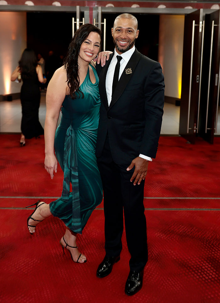 Woman in a body-hugging green dress showing her figure, standing next to a man in a black suit on a red carpet.