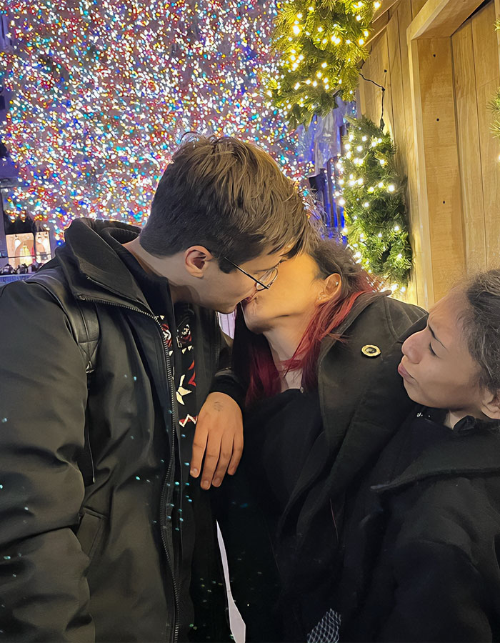 Conjoined twin Carmen Andrade kissing her husband under festive lights, with a third person watching nearby.