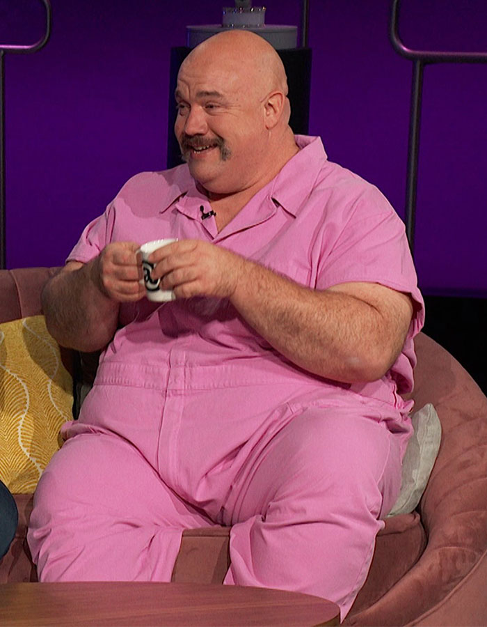 Plus-size man in pink outfit smiling and holding a cup during a casual interview on a purple set background. Plus-size man in pink outfit smiling and holding a cup during a casual interview on a purple set background.