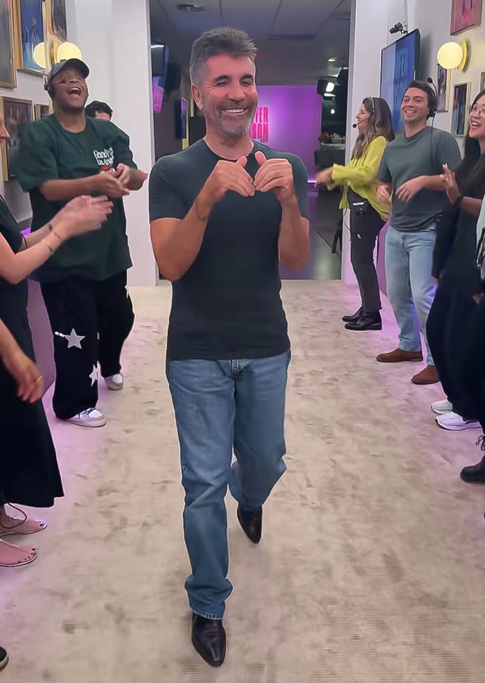 Simon Cowell smiling and walking confidently with a group of people clapping and smiling indoors after a bike accident.