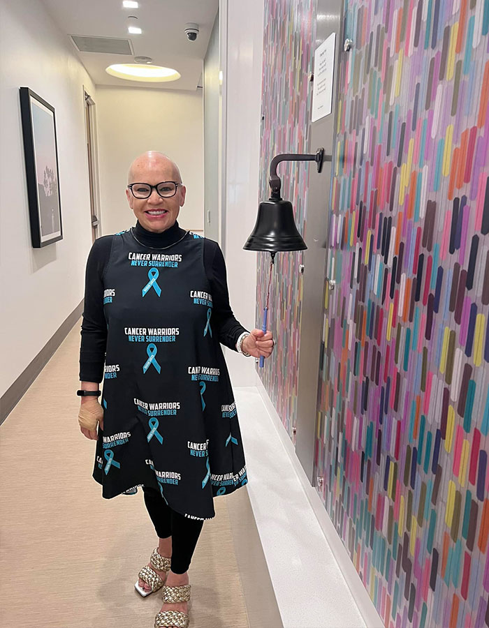 Woman in cancer warrior dress ringing bell after completing cancer treatment, smiling in a medical hallway.