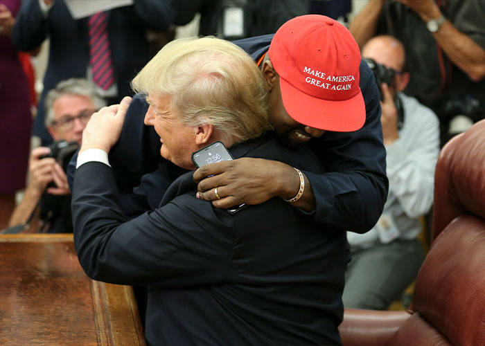 Kanye West wearing red hat hugging former president in a public setting with photographers capturing the moment.