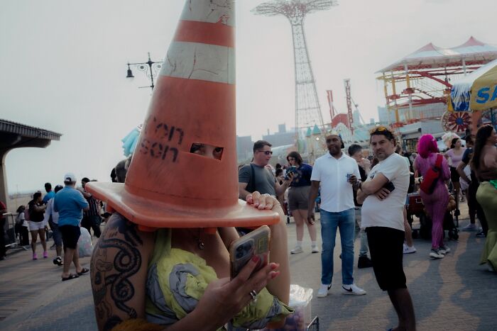 Person wearing a large traffic cone on their head surrounded by people at a busy outdoor event in a striking street photo.