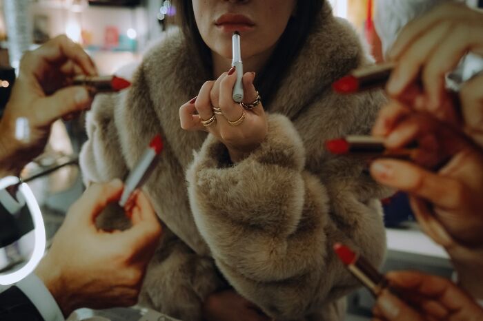 Striking street photo of a woman in a fur coat holding a cigarette while multiple hands apply red lipstick around her.