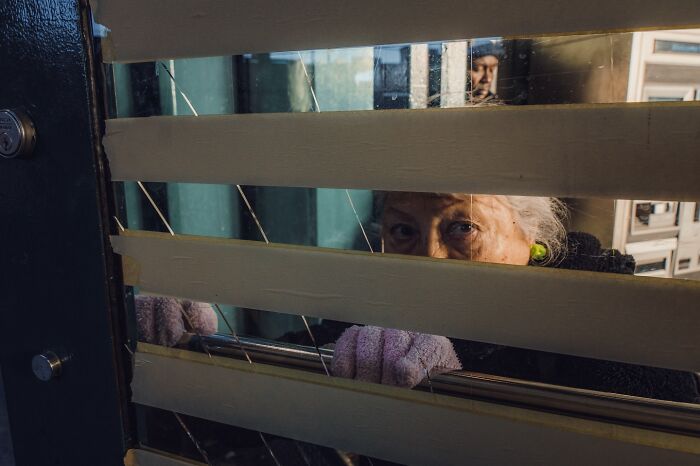Elderly woman peering through window blinds in a striking street photo with cinematic lighting and mood.
