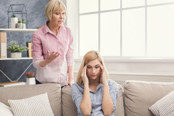 Woman sitting on a couch with headache as older woman stands behind her showing frustration in a bright living room.