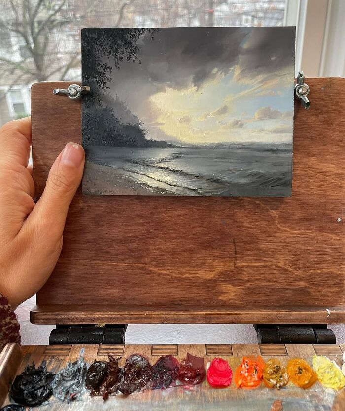 Hand holding a detailed painting by Dina Brodsky showing a serene natural world scene with a beach and cloudy sky.