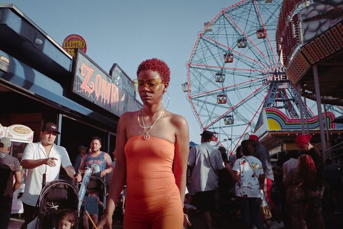 Woman in an orange outfit walking through a crowded amusement park, captured in striking street photography style.