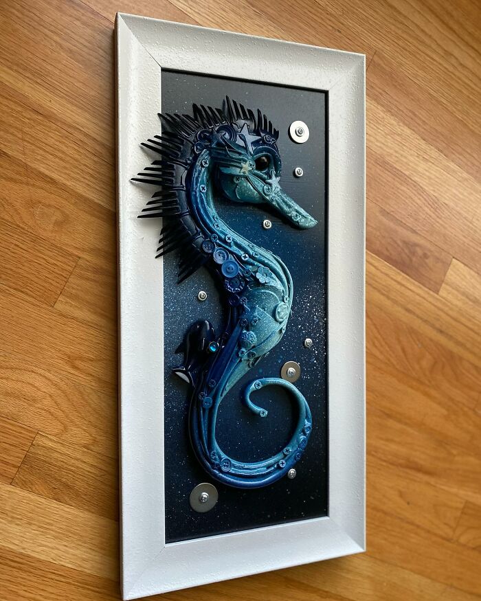 Blue seahorse sculpture made from trash and scrap materials, displayed in a white frame on a wooden floor.