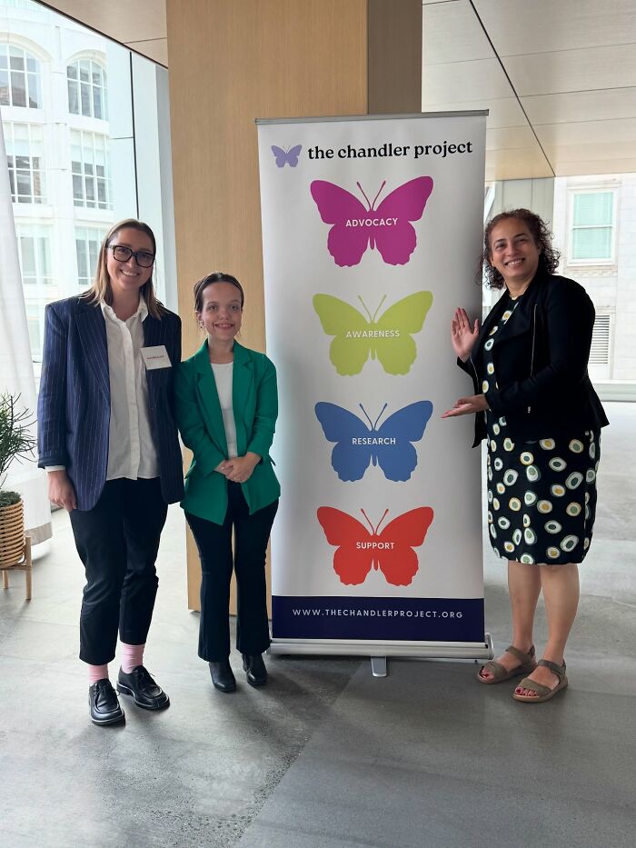 31YO woman with dwarfism standing beside advocacy banner with two other women at an indoor event.