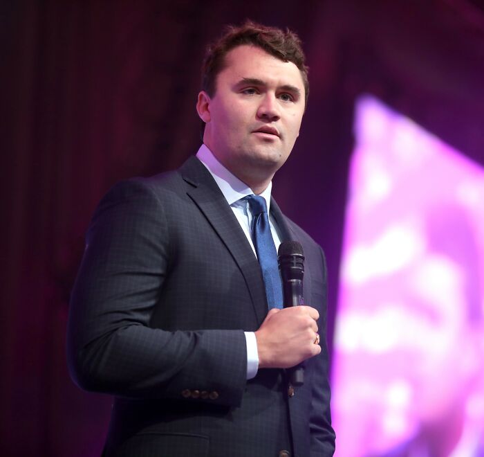 Man in a dark suit holding a microphone, speaking at an event with purple and pink blurred lights in the background.
