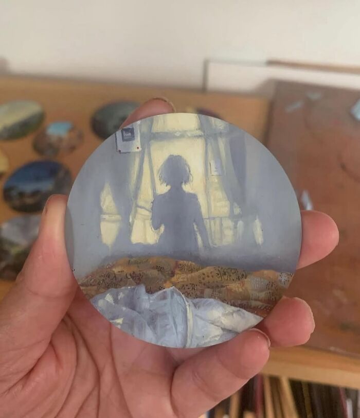 Hand holding a small round painting by Dina Brodsky showing a shadowed figure by a window in soft natural light.