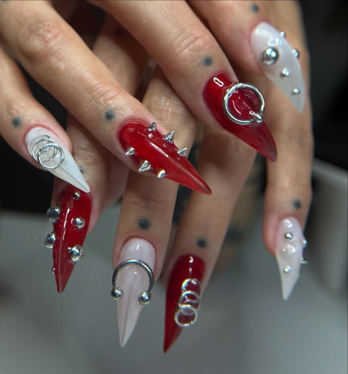 Red and white edgy nail designs featuring silver spikes, rings, and studs on long stiletto-shaped nails breaking all the rules.
