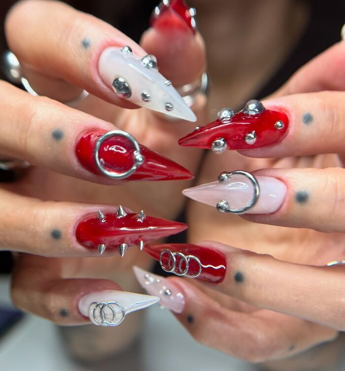 Red and white edgy nail designs featuring silver spikes, rings, and studs on long stiletto-shaped nails breaking all the rules.
