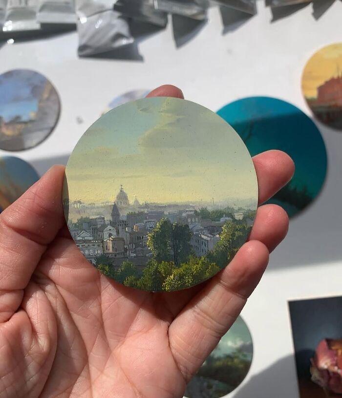 Hand holding a detailed painting by Dina Brodsky depicting a scenic view of trees and buildings at sunset.
