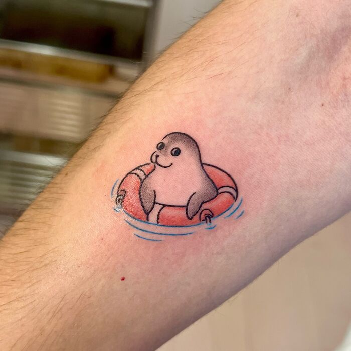 Cute seal tattoo in a lifesaver ring with water details, an adorable animal tattoo by Korean artist Buoy on forearm.