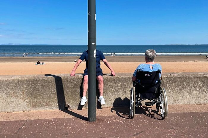Man sitting behind street pole blocking his face and person in wheelchair beside him on beach promenade in street photo