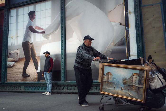 Man pushing cart with a framed painting on a street featuring large advertisement resembling movie stills.