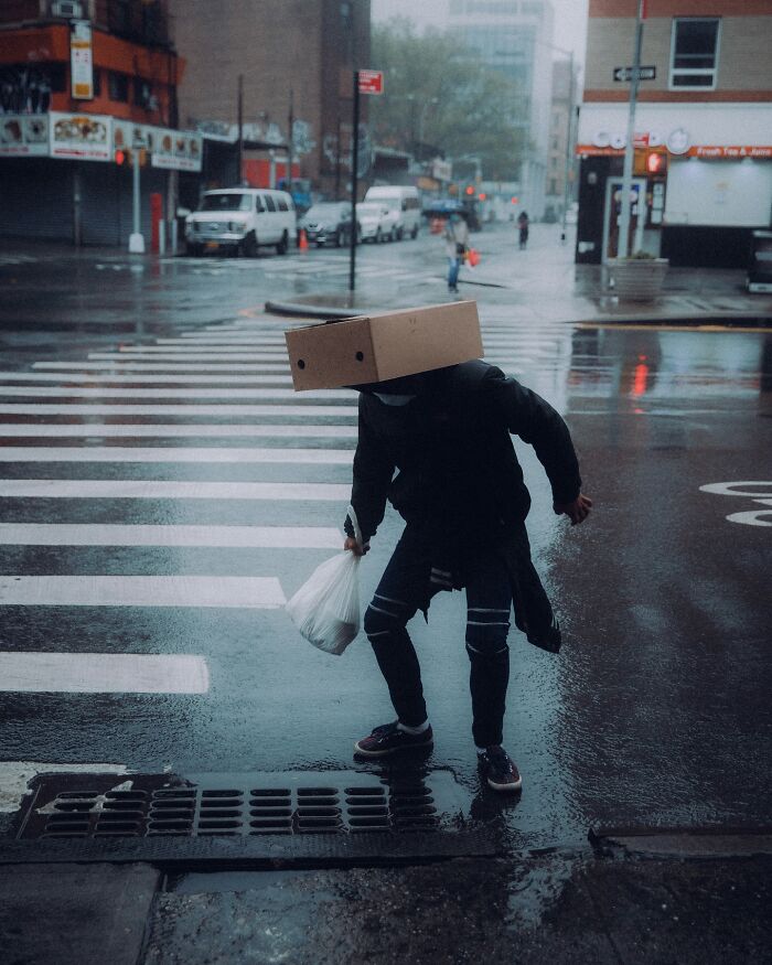 Person wearing a cardboard box on their head crossing a rainy street, capturing striking street photos with a cinematic feel.