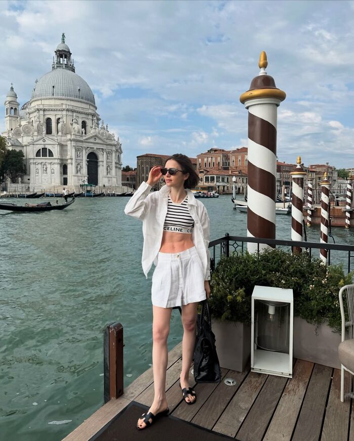 Lily Collins with extreme thinness, wearing white outfit and sunglasses, standing by Venice canal and historic architecture.