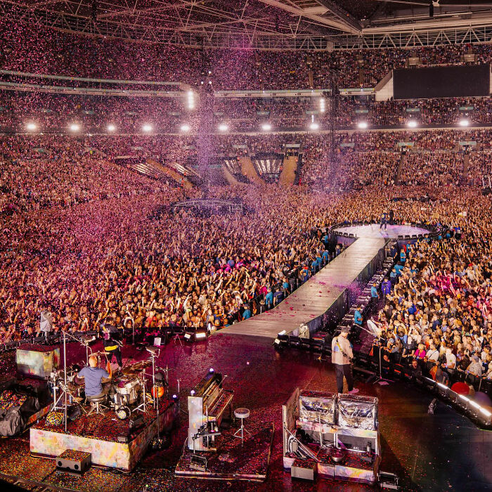Coldplay performing on a large stage with a massive crowd at a stadium concert amid confetti effects.