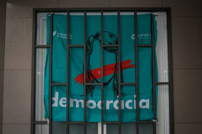 A street photography image showing a teal banner with a red stripe over a face behind barred window bars.
