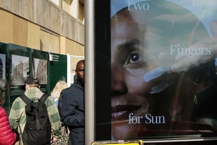 Street photo showing pedestrians near a reflective advertisement with a close-up face and text in an urban setting.