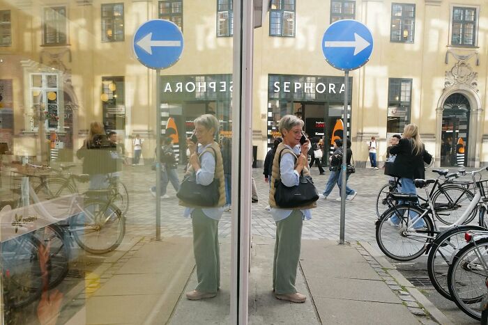 Street photo showing a woman taking a selfie reflected in glass with traffic signs and bicycles in an urban setting.