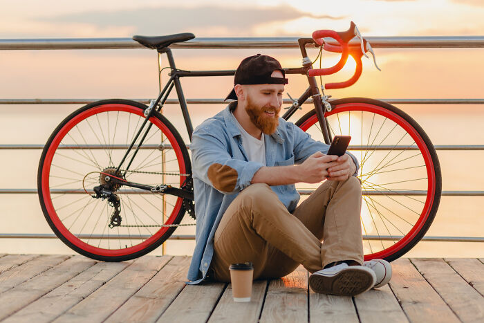 Man sitting by a bike on a wooden pier, surfing online and discovering his partner's secret stories on a phone.
