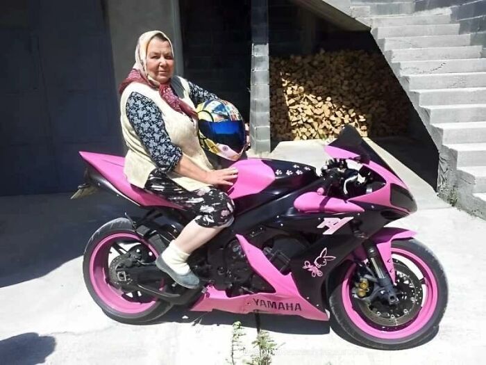 Elderly woman in traditional Eastern European attire sitting on a pink Yamaha motorcycle holding a helmet.