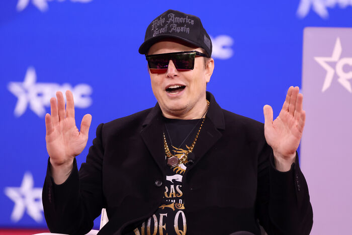 Elon Musk wearing sunglasses and a black cap, gesturing with raised hands during a public event discussion.