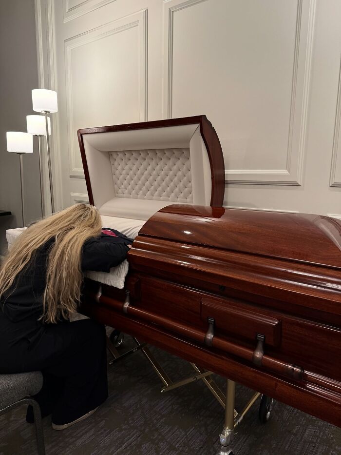 Woman with long blonde hair grieving beside a polished wooden casket during a funeral amid heated feminist movement debate.