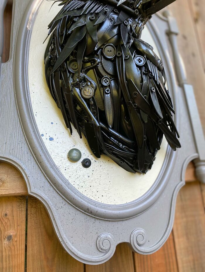 Sculpture of a bird made from repurposed metal trash pieces displayed on a decorative frame against a wooden background.