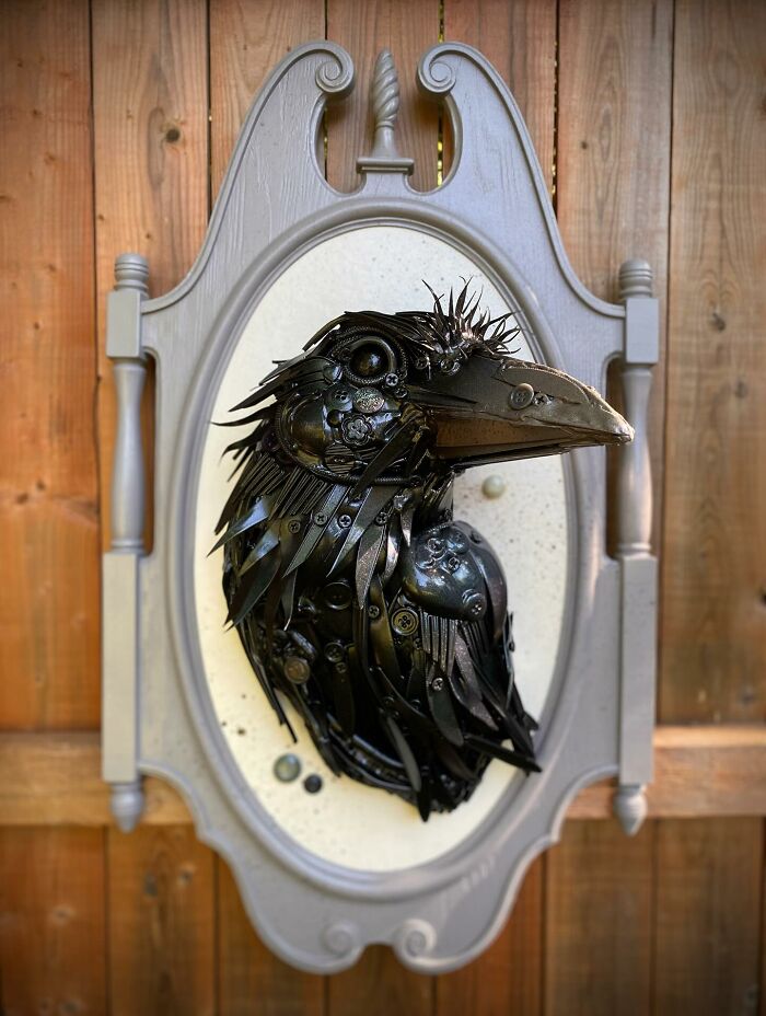 Sculpture of a bird made from repurposed metal trash pieces displayed on a decorative frame against a wooden background.