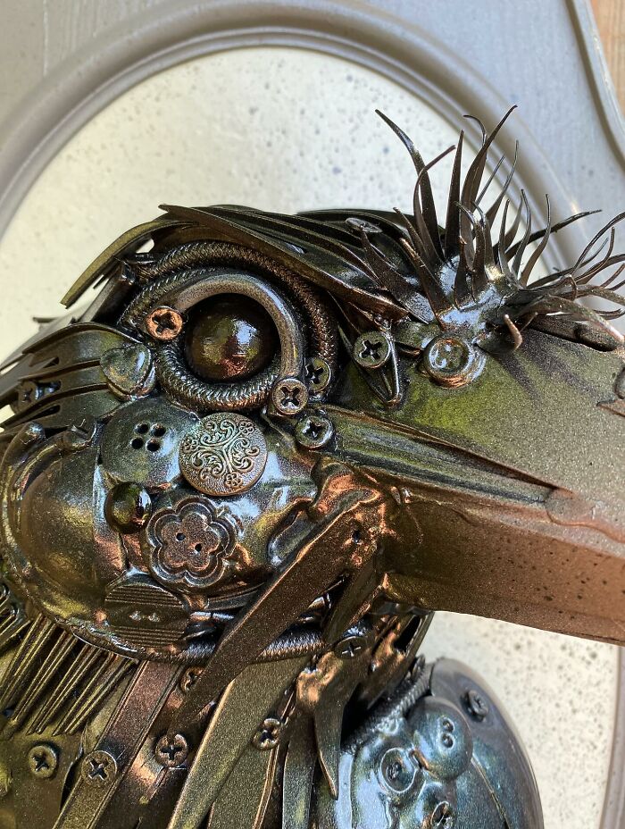 Sculpture of a bird made from repurposed metal trash pieces displayed on a decorative frame against a wooden background.