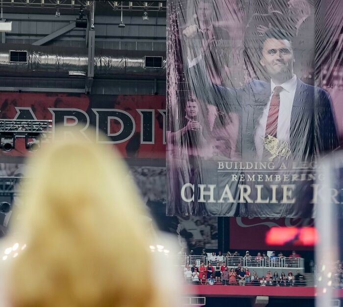 Woman with blonde hair watching a large poster of Charlie Kirk at a memorial event with crowd in the background.