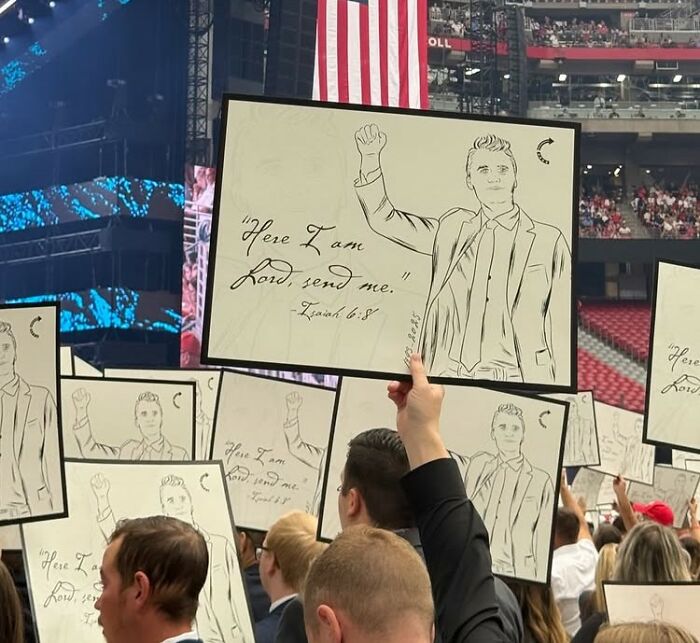 Crowd holding signs with raised fists at Charlie Kirk's funeral, with mass fury compared to historic events.