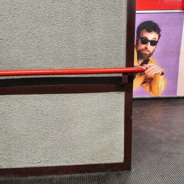 Man in sunglasses from a poster appears to hold a red handrail, creating a funny street photo coincidence.