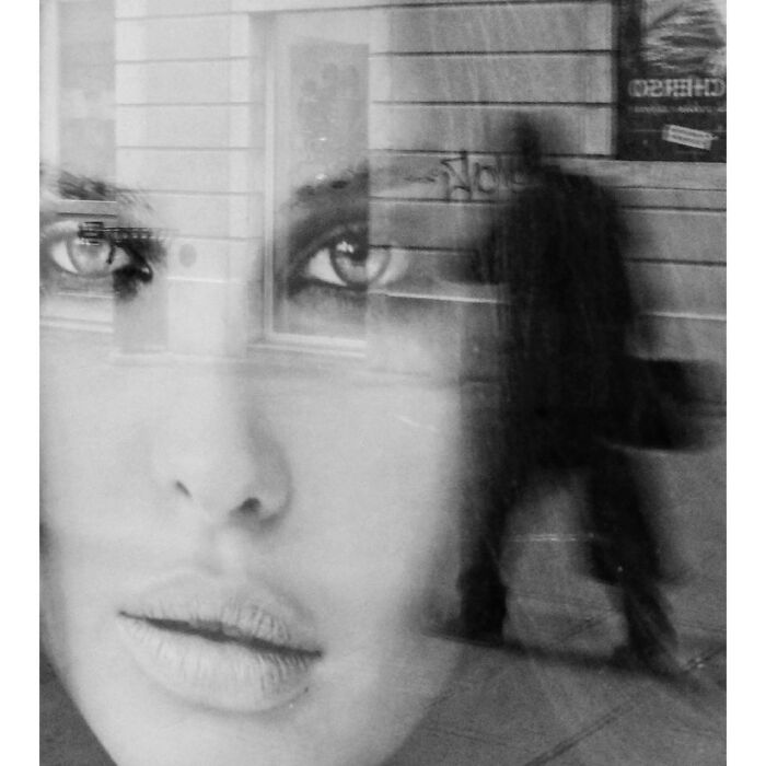 Black and white street photo showing a layered reflection of a woman's face and a walking figure, capturing funny coincidences.