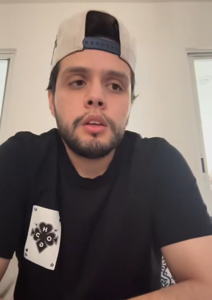 Young man wearing a black shirt and backward cap, appearing thoughtful in a bright indoor setting.